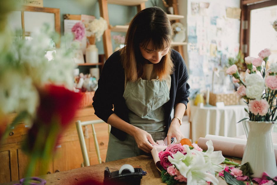 A female florist in a floral shop is arranging a bouquet of fresh flowers, with pink roses, white lilies, and other blooms carefully wrapped in pink paper. She is wearing a light gray apron over a dar