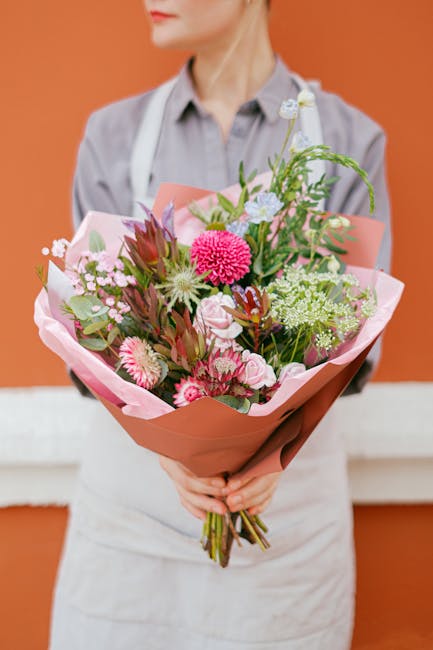 A person wearing a light gray shirt and white apron holds a vibrant bouquet of fresh flowers against a warm orange background. The bouquet features a variety of blooms including pink gerbera daisies, 