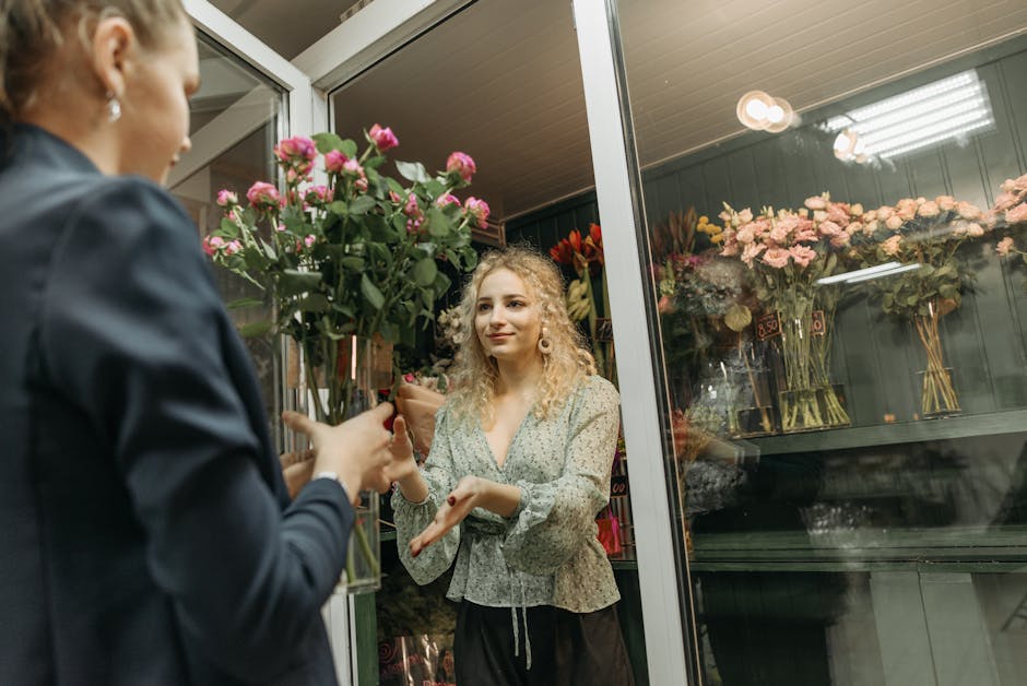 A young woman with curly blonde hair and fair skin is inside a flower shop, accepting a bouquet of fresh pink roses wrapped in brown paper from a florist. The bouquet is arranged with lush green leave