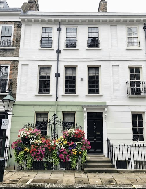 A flowering display in front of a white, three-story residential building with black-framed windows and a black door. The arrangement features a variety of fresh flowers, including pink, white, yellow