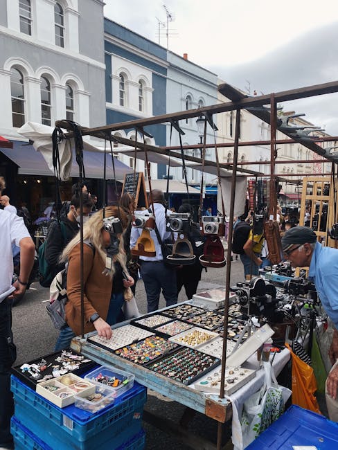 A busy outdoor market scene on Portobello Road with a floral and jewelry stall in the foreground. The stall features a variety of accessories displayed on a table, including colorful jewelry, brooches