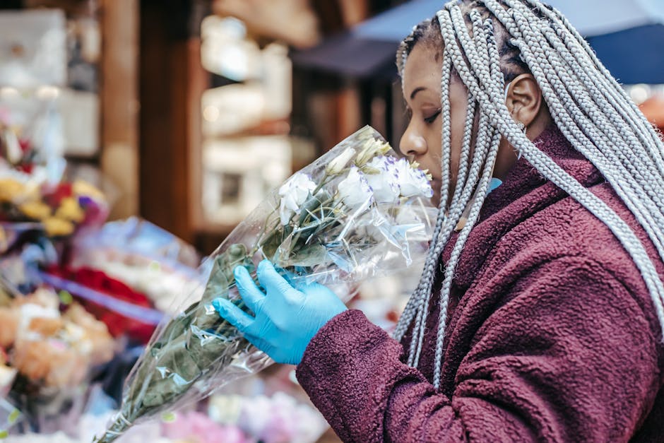 A woman at Portobello Road Market, wearing a maroon fleece jacket and light gray braids, is holding a bouquet of fresh white roses wrapped in clear cellophane. She is inspecting the flowers closely, w