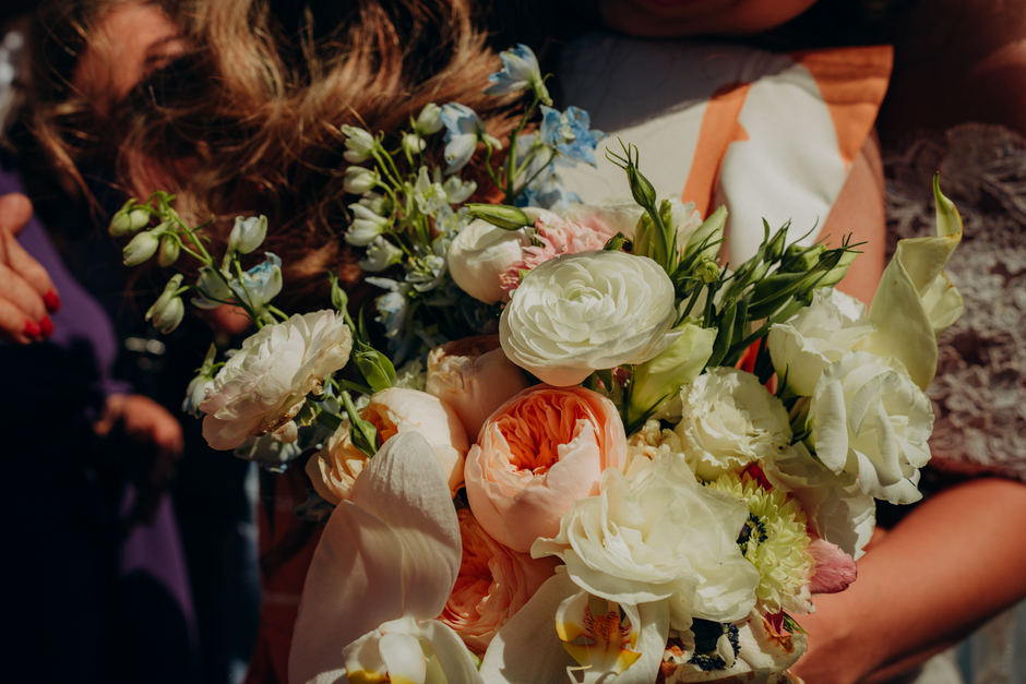 A close-up view of a bouquet featuring a mix of fresh flowers including white ranunculus, cream calla lilies, pale peach peonies, and light blue delphiniums, arranged with green foliage. The flowers a