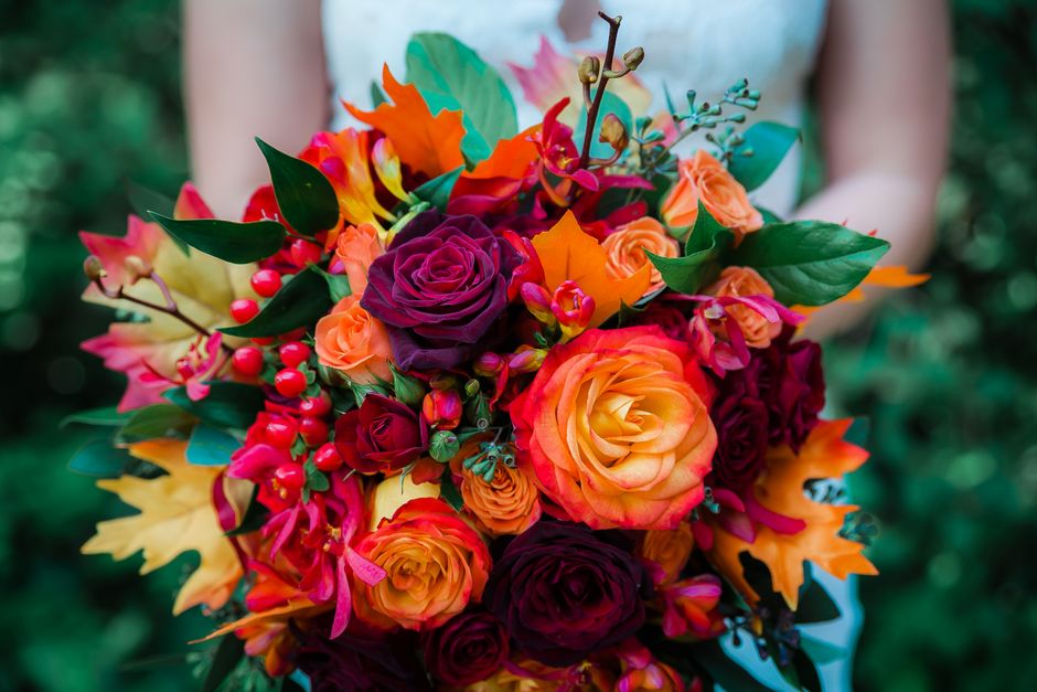 A vibrant floral bouquet featuring a mix of roses in shades of orange, yellow, deep purple, and burgundy, interspersed with bright red berries, pink azaleas, and lush green leaves. The arrangement is