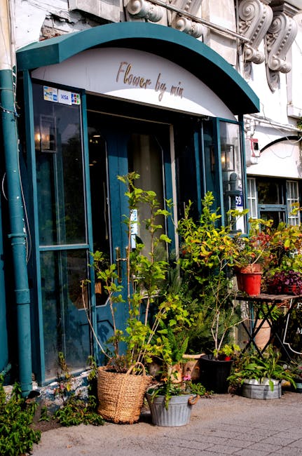 The storefront of 'Flower by Mina' features a teal-colored awning with elegant white lettering, situated above a glass door with dark teal framing. In front of the shop, there are lush green potted pl