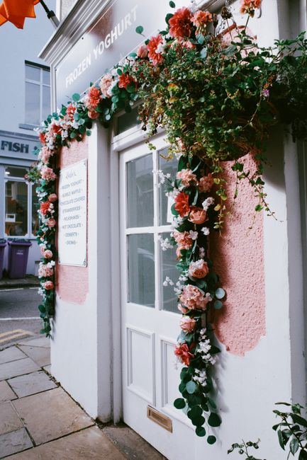 A floral arrangement framing a white, wooden door with a small glass window, set against a pink-painted exterior wall. The arrangement features an abundant garland of roses in shades of pink and coral